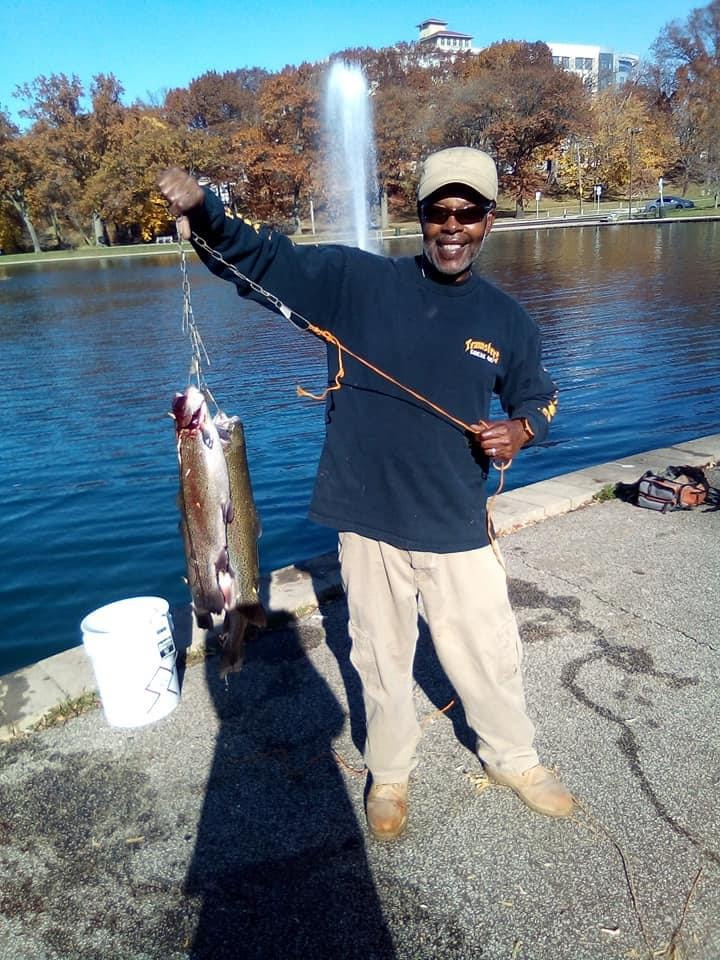 Black man wearing a tan hat, long sleeved navy shirt and tan pants holds up trout caught at Rockefeller Lagoon.